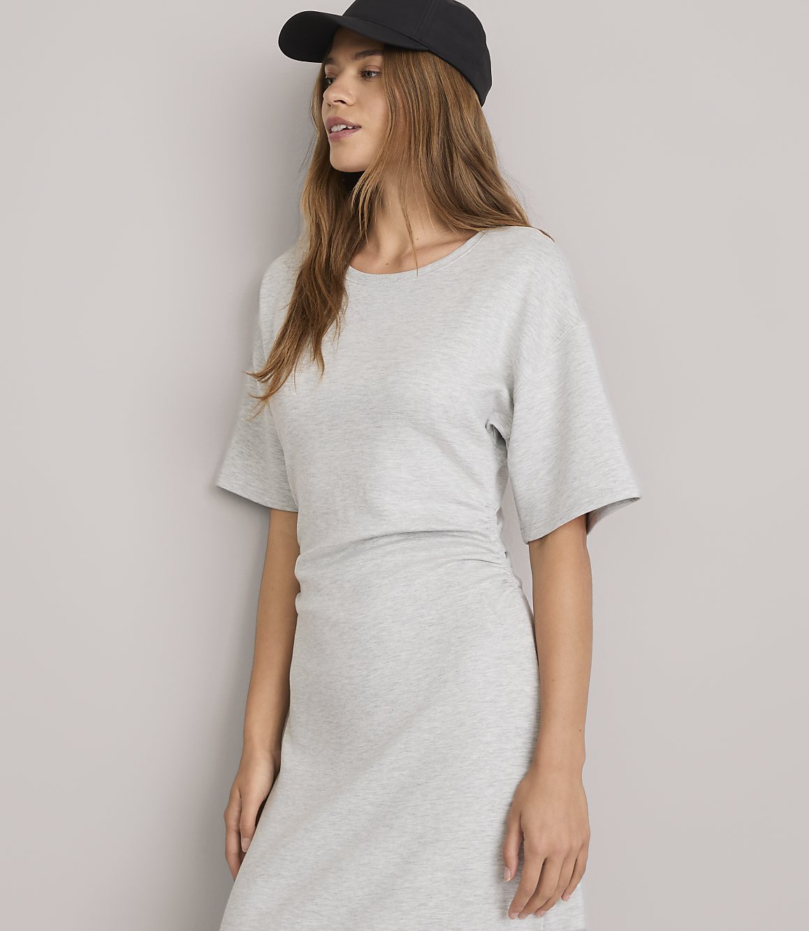 A young woman in a grey dress and black baseball cap stands against a plain grey background.