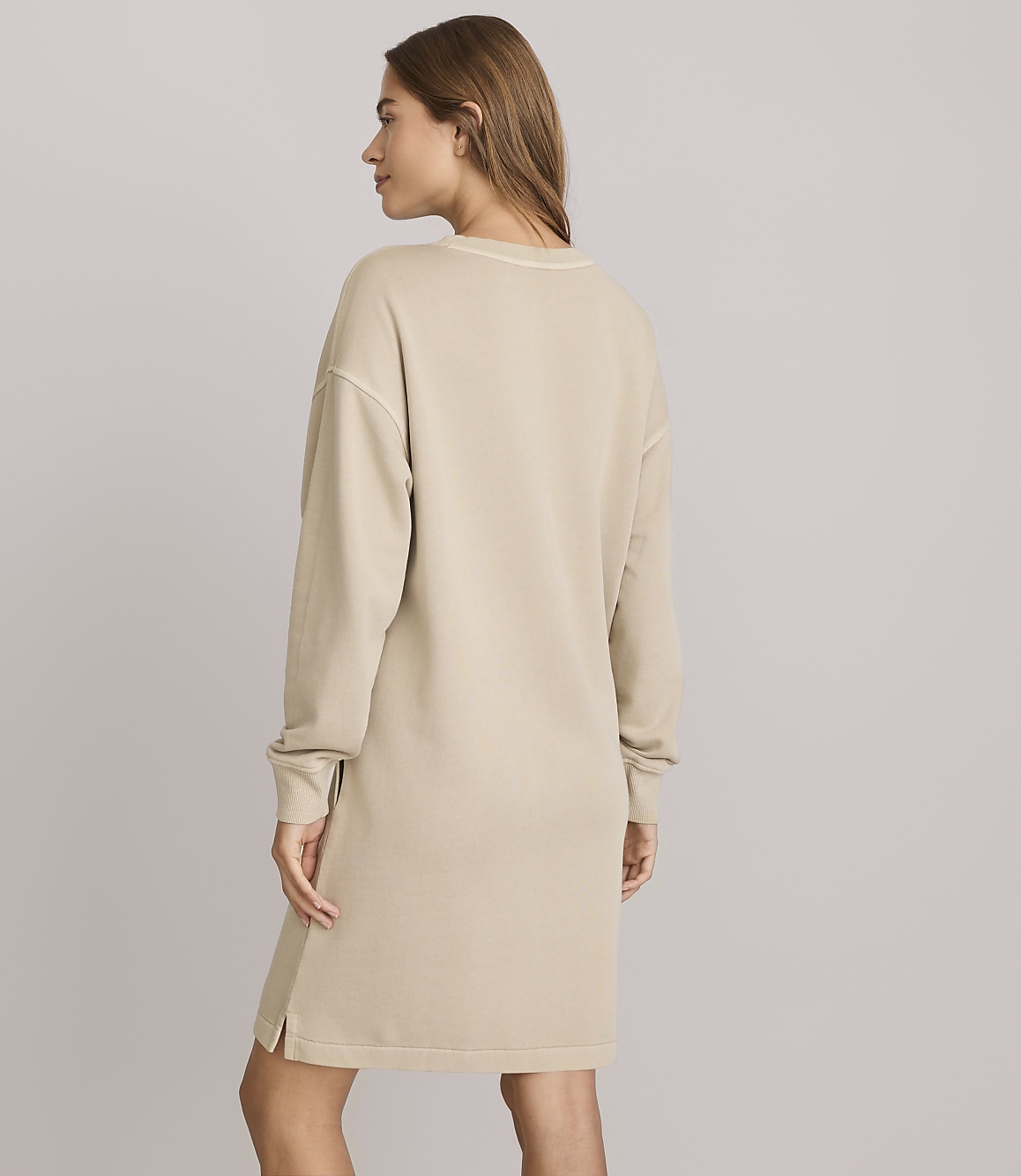 A woman in a beige sweatshirt dress stands against a plain, light gray background.