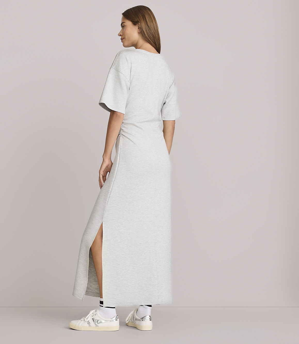A woman in a grey maxi dress and sneakers stands against a plain grey background.