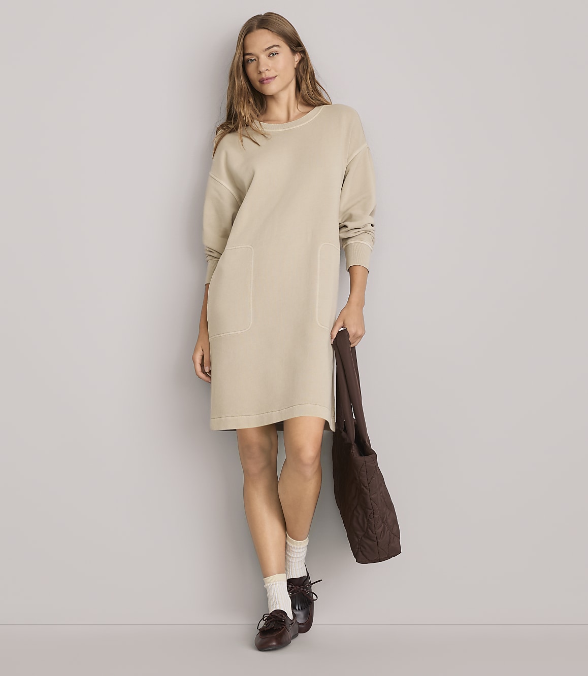 A woman in a beige dress and loafers stands against a plain gray background, holding a brown tote bag.