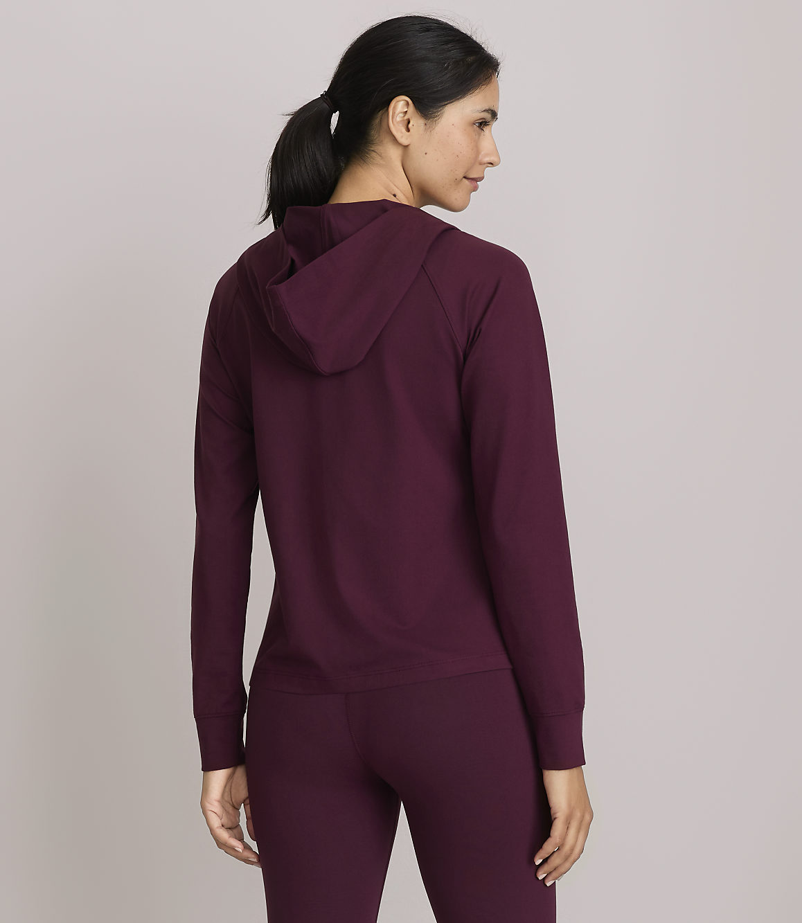 A woman in a burgundy hoodie and leggings stands facing away from the camera against a plain gray background.