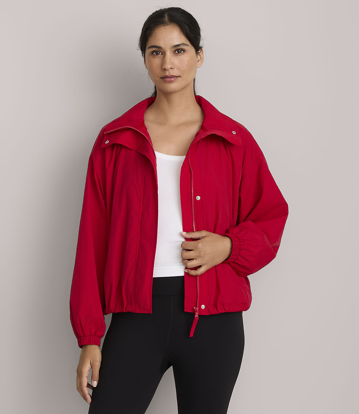 A woman in a red jacket and white tank top stands against a plain gray background.
