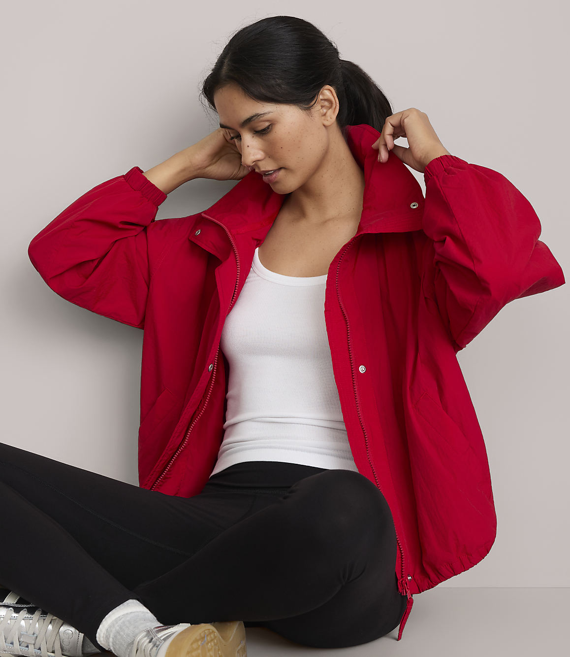 A woman in a red jacket and black leggings sits against a plain gray background.