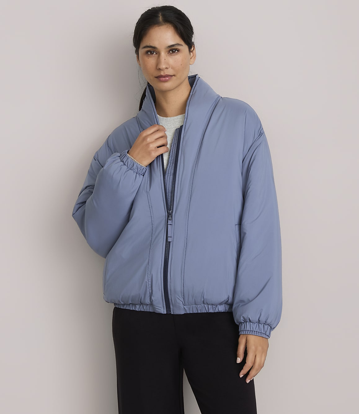 A woman in a blue puffer jacket stands against a plain gray background.