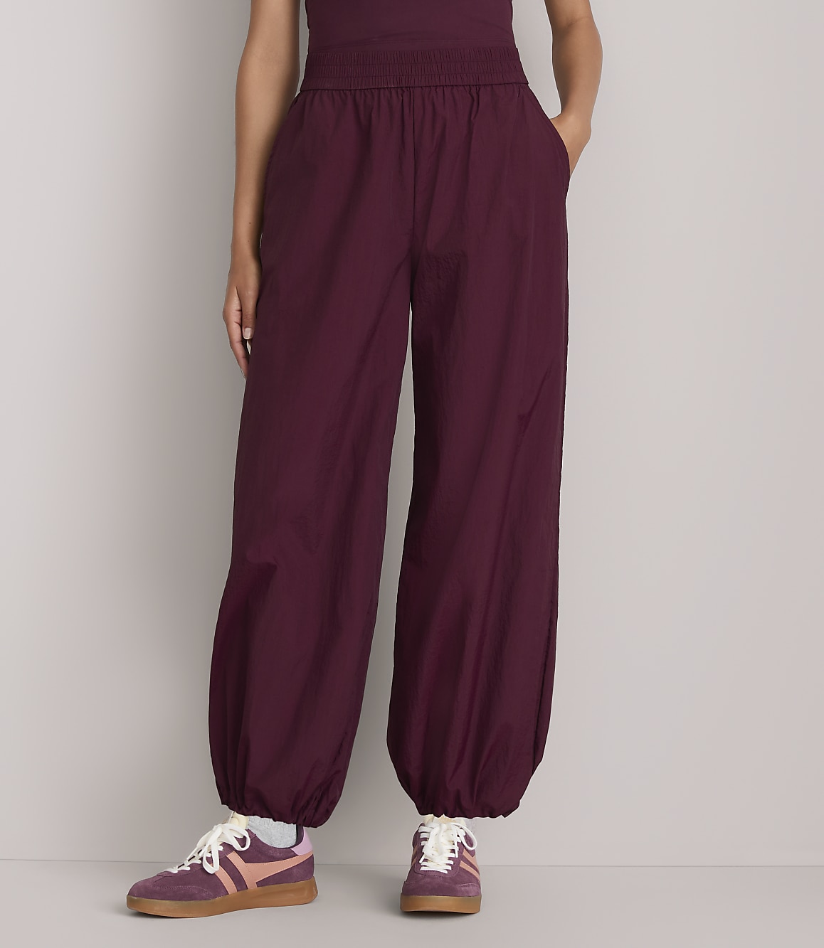 A person stands in loose-fitting burgundy pants with elastic cuffs and sneakers against a plain gray background.