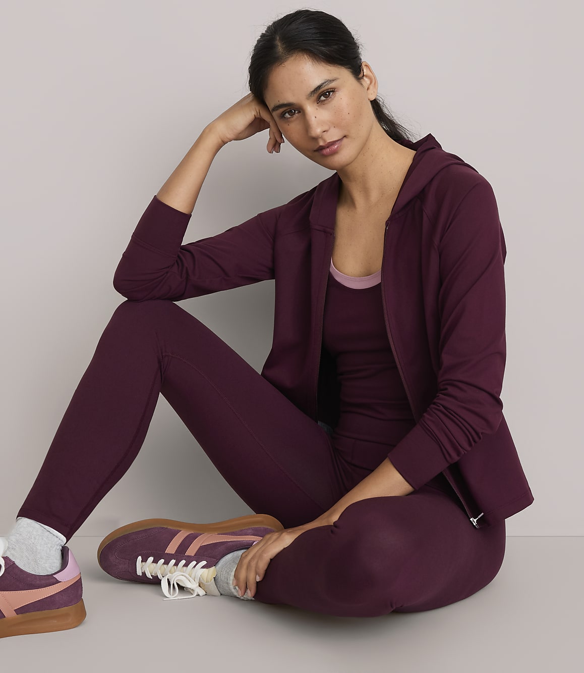 A woman in burgundy activewear sits on a plain gray floor with a matching hoodie and leggings, her hand resting on her head.