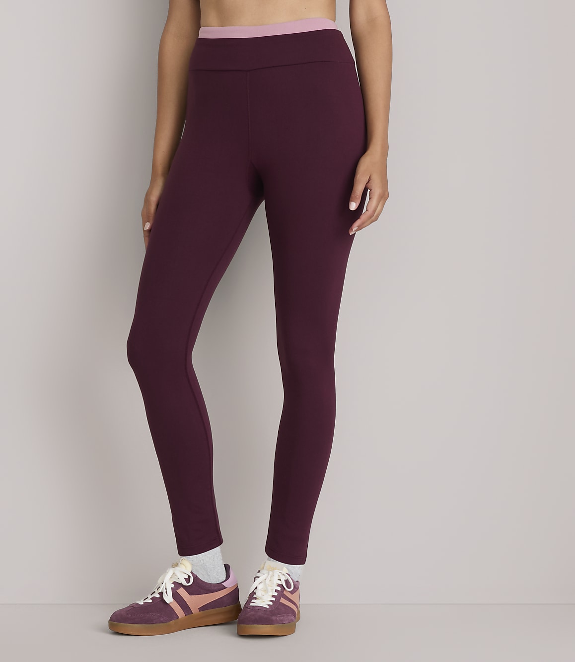 A person wearing burgundy leggings and pink and brown sneakers stands against a plain gray background.