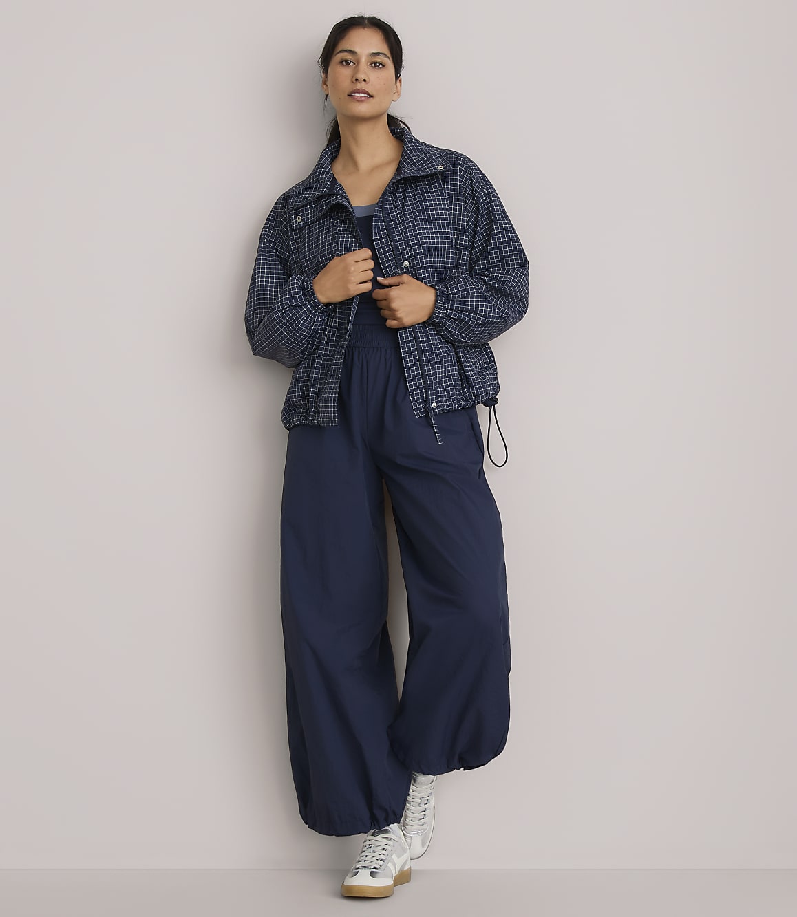 A woman in a patterned jacket and wide-leg pants stands against a plain, light-colored wall.