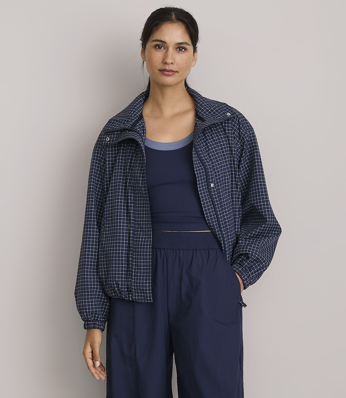 A woman in a blue checkered jacket and navy blue athletic wear stands against a plain gray background.