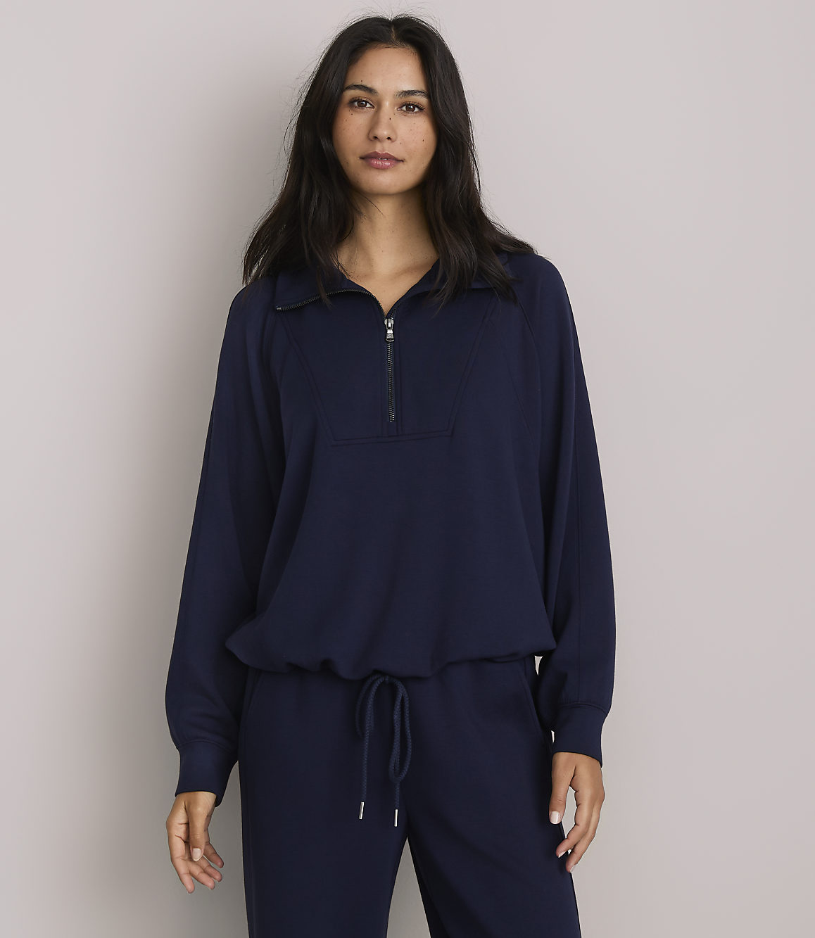 A woman in a navy blue quarter-zip sweatshirt and matching pants stands against a plain gray background.