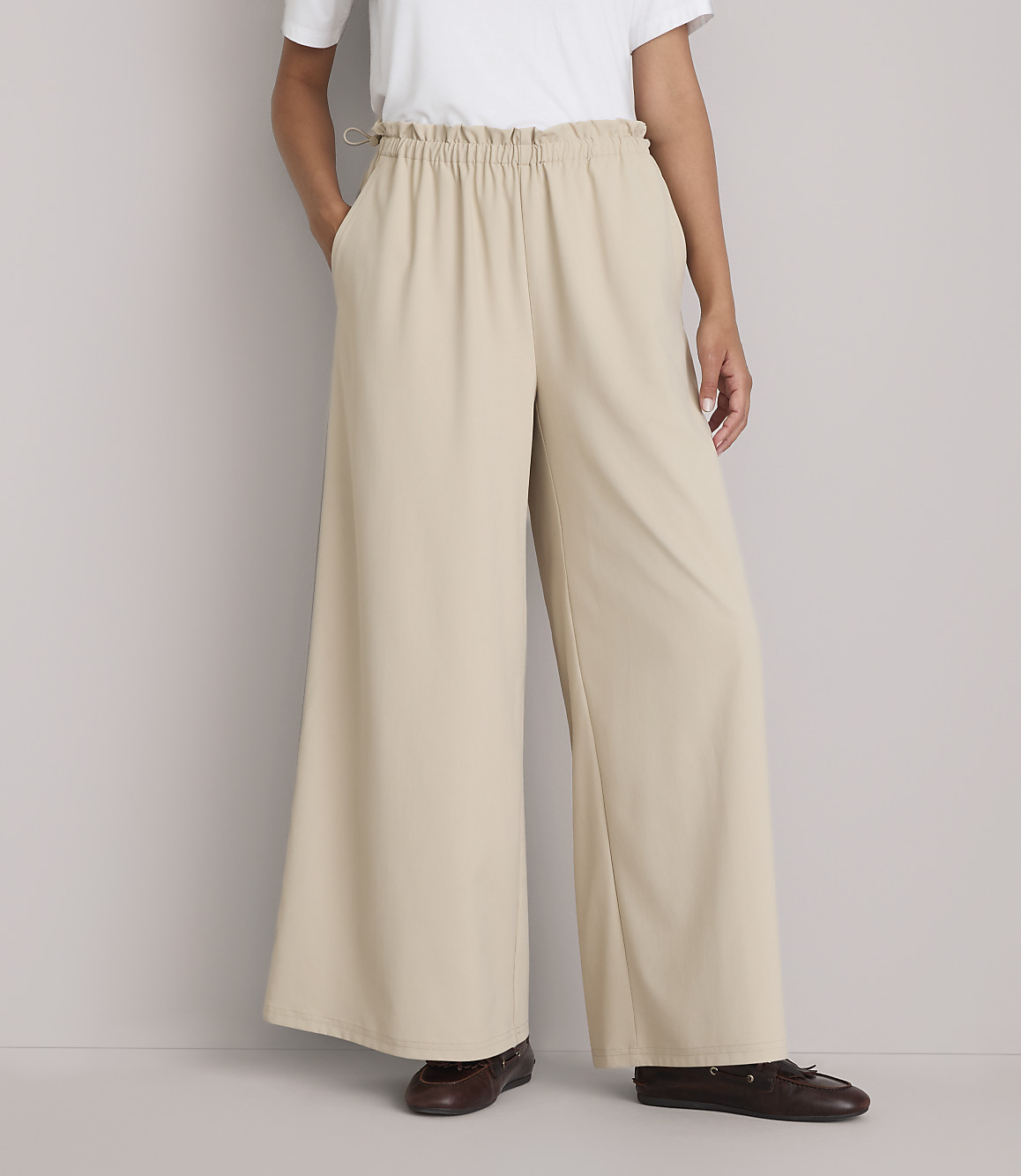 A person stands in wide-leg beige pants and brown shoes against a plain gray background.