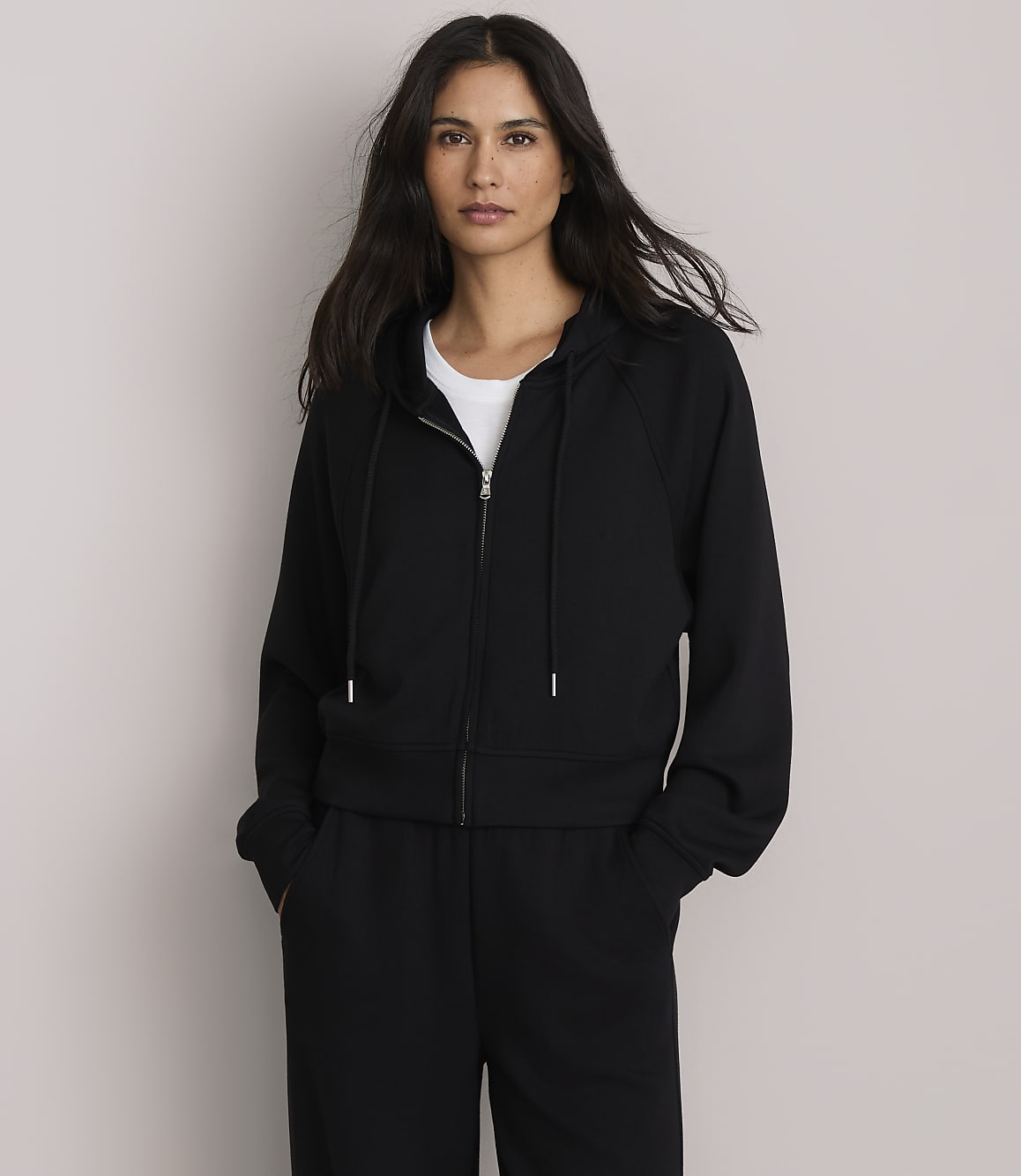 A woman in a black zip-up hoodie and sweatpants stands against a plain, light gray background.
