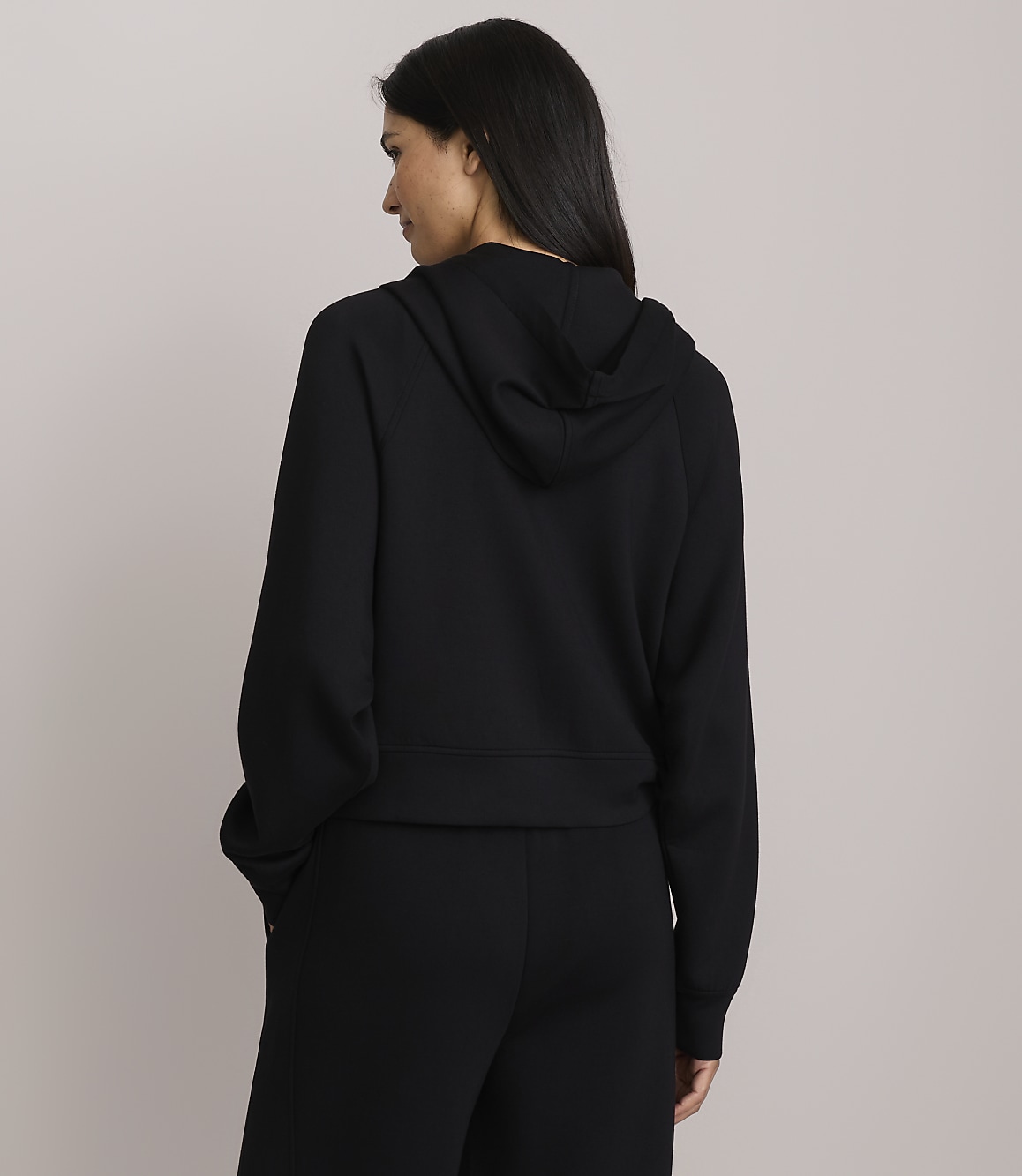 A woman in a black hoodie and pants stands with her back to the camera against a plain gray background.
