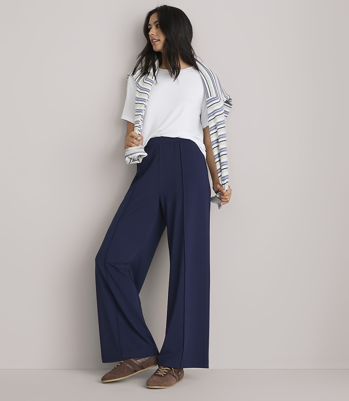 A woman in a white t-shirt and navy wide-leg pants stands against a plain gray background, with a striped sweater draped over her shoulders.
