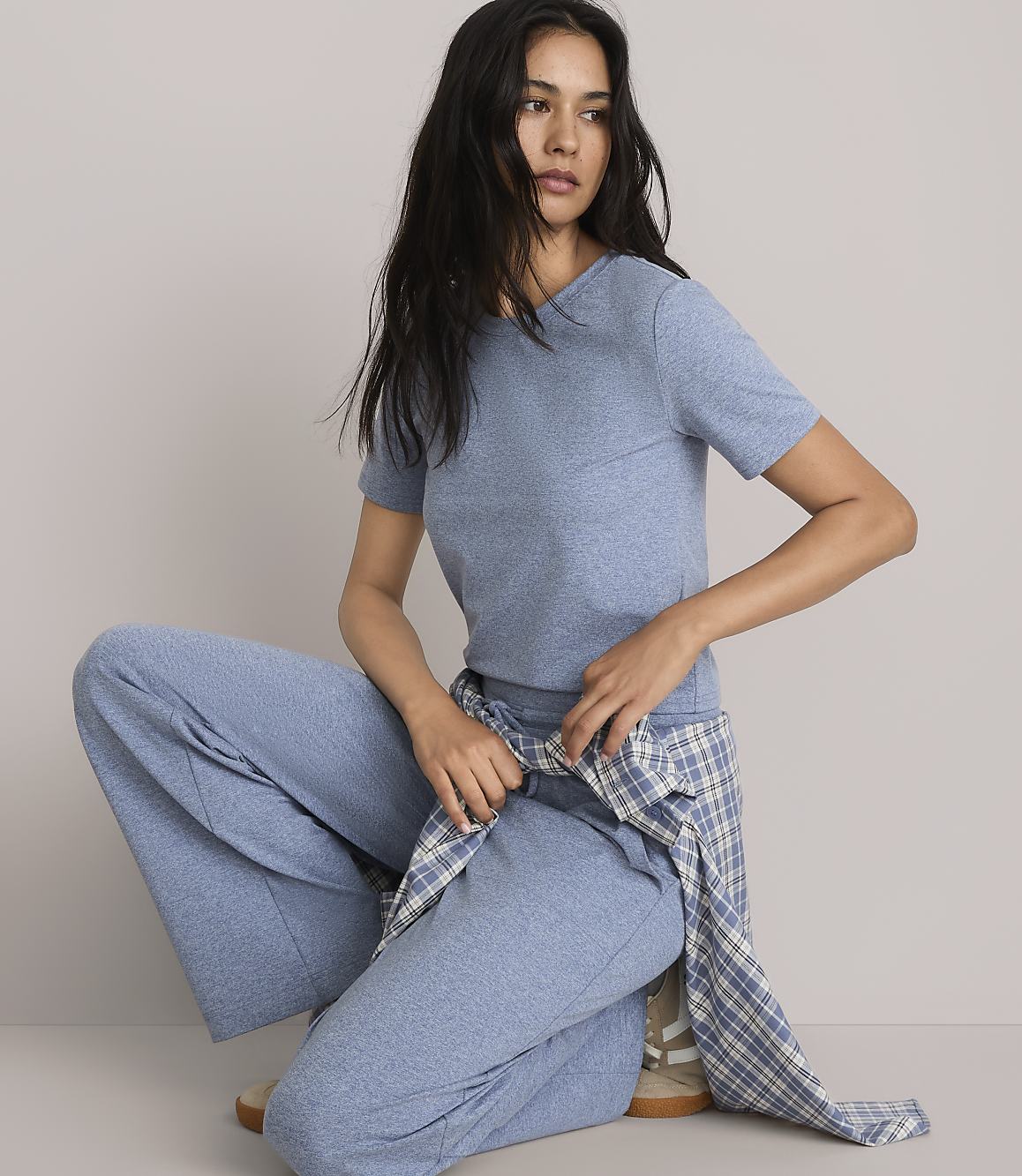 A woman in a blue t-shirt and wide-leg pants poses against a plain gray background, with a plaid shirt tied around her waist.