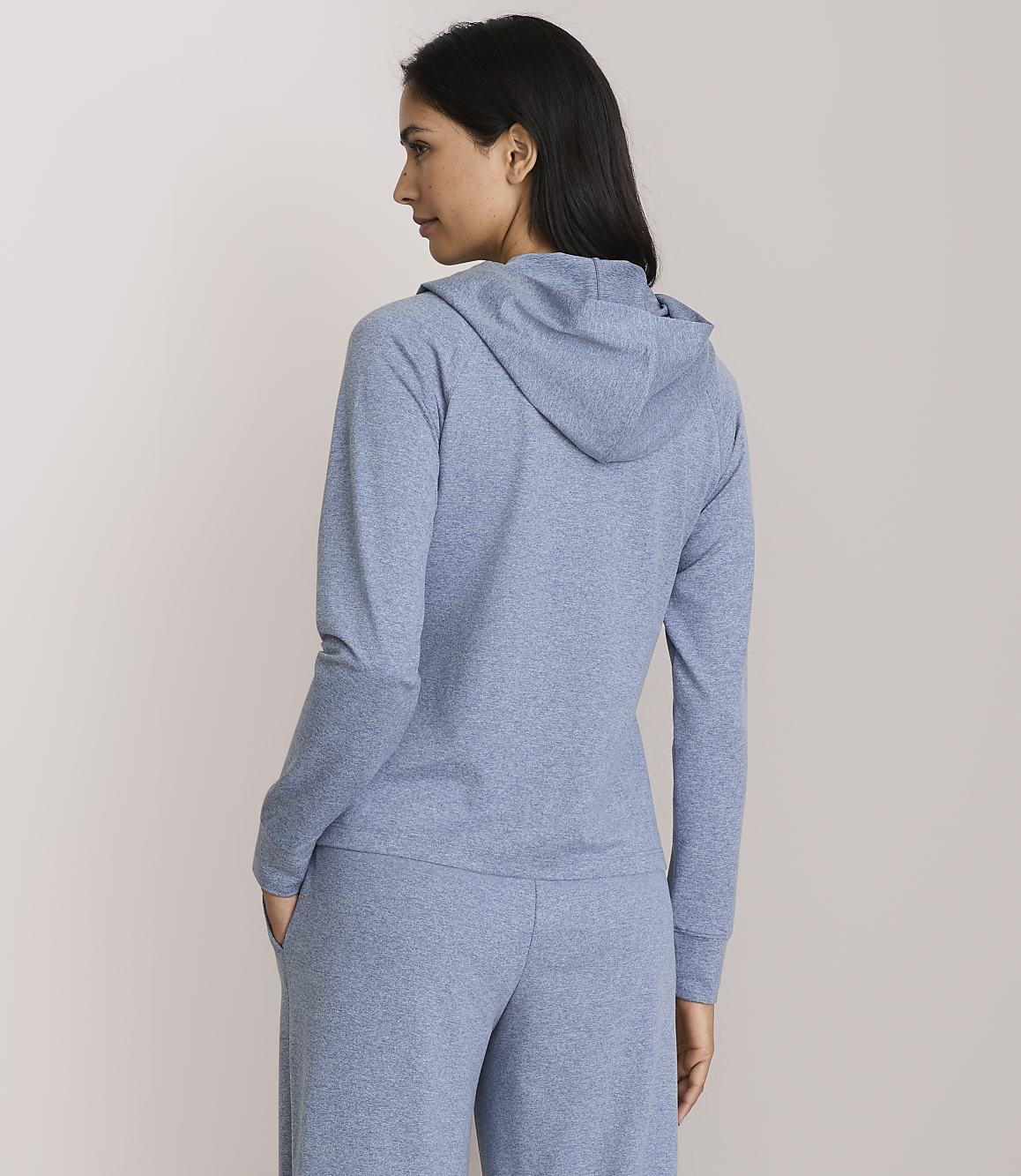 A woman in a blue hoodie and pants stands against a plain, light pink background.