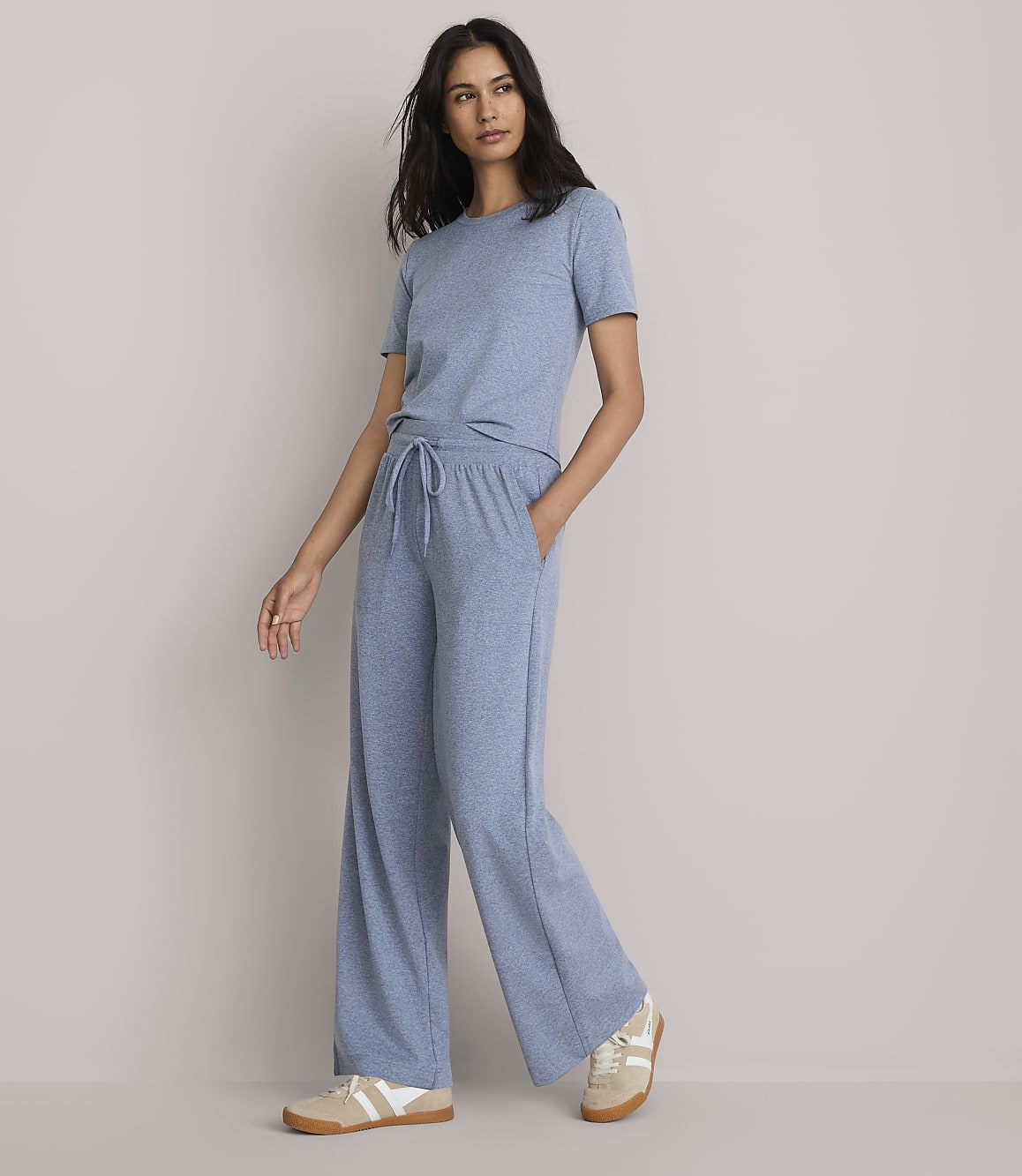 A woman in a blue t-shirt and wide-leg pants stands against a plain, light gray background.