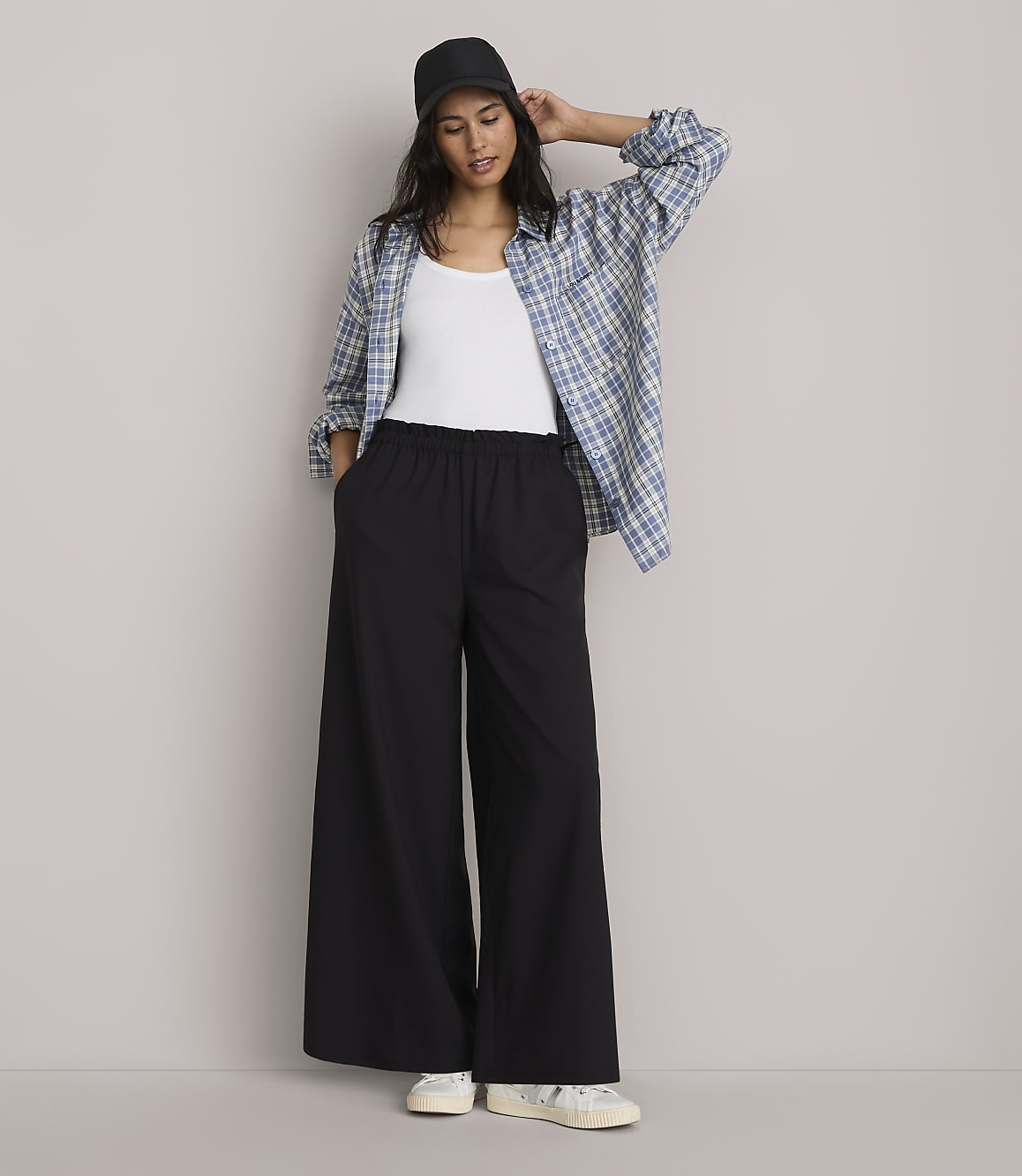 A woman in a white tank top, black wide-leg pants, and an open plaid shirt stands against a plain gray background.