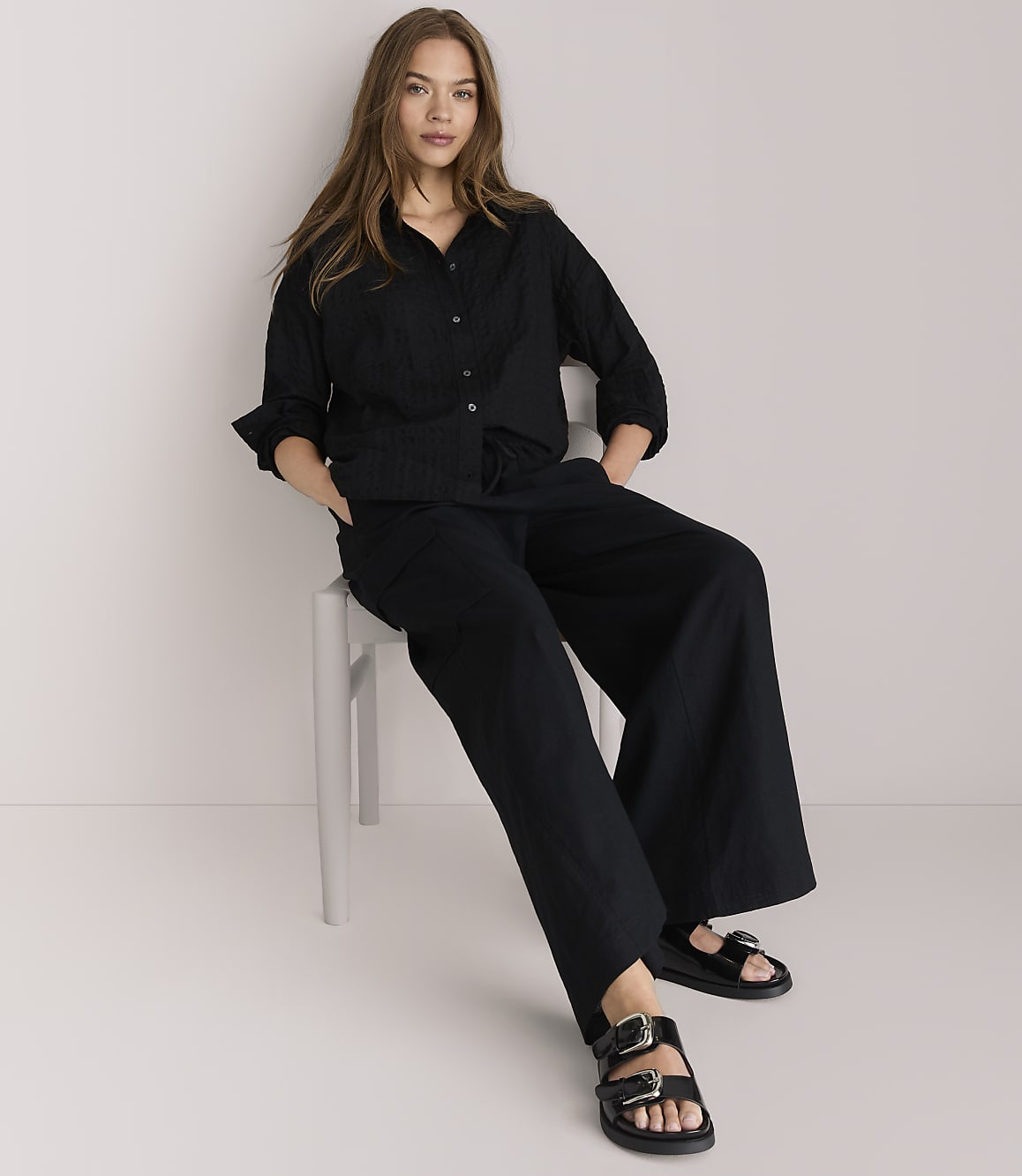 A woman in a black textured shirt and black pants stands against a plain white background.