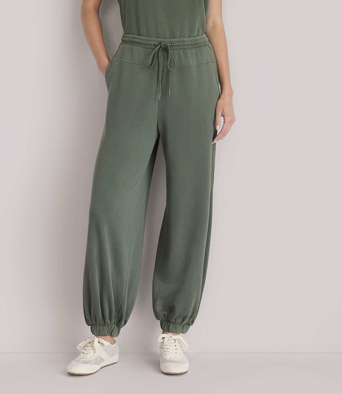 A person wearing olive green sweatpants and sneakers stands against a plain white background.