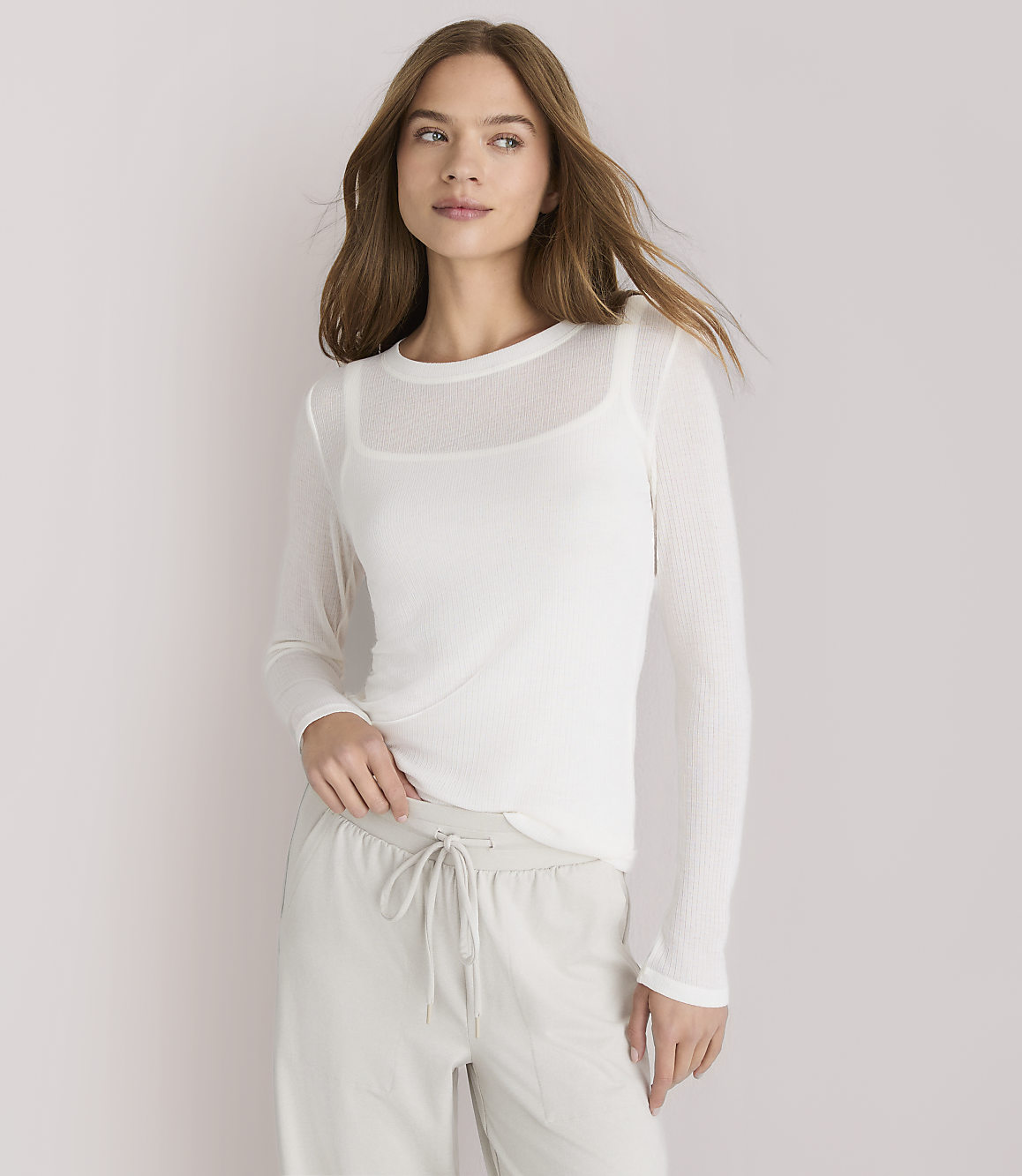 A woman in a white sheer long-sleeved top and light gray drawstring pants stands against a plain white background.