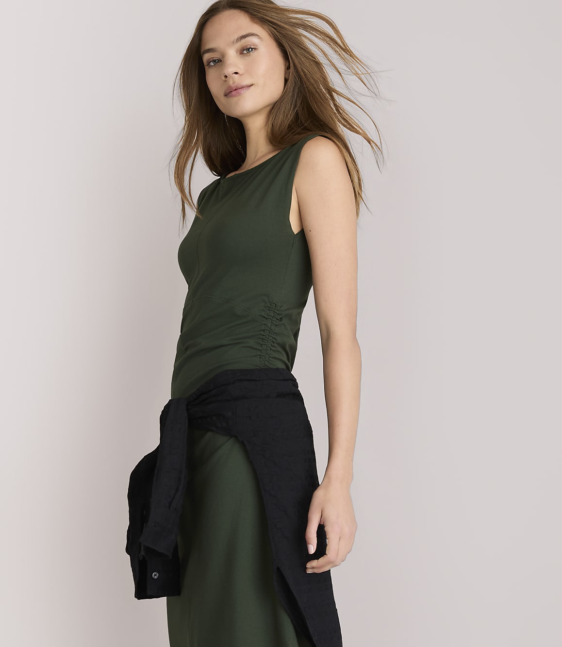 A woman in a green dress with a black sweater tied around her waist stands against a plain white background.