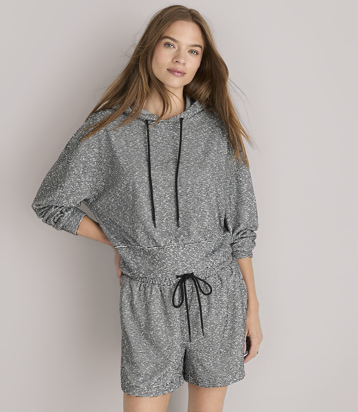 A woman in a grey patterned hoodie and shorts sits against a plain grey background.