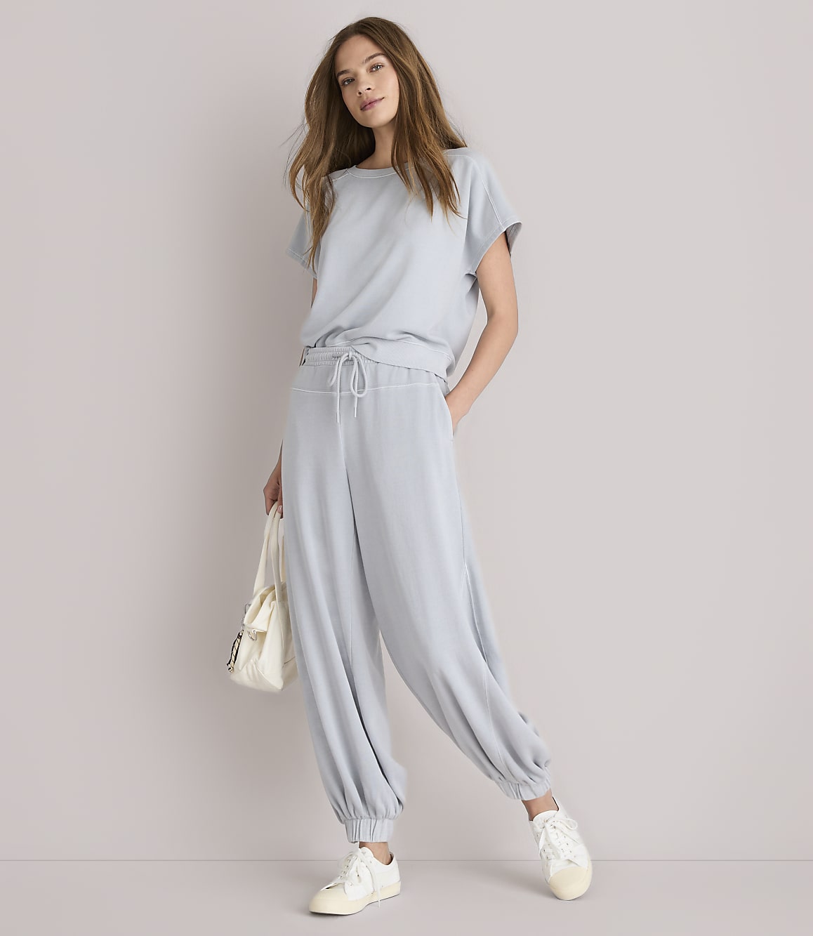 A woman in a light blue loungewear set and white sneakers stands against a plain white background, holding a cream-colored tote bag.