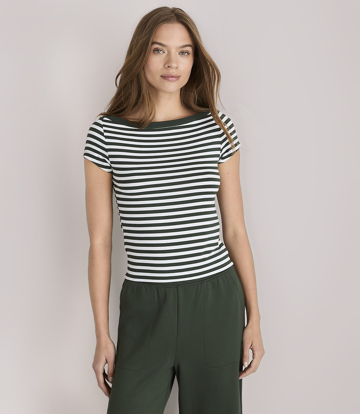A woman in a striped shirt and green pants stands against a plain white background.