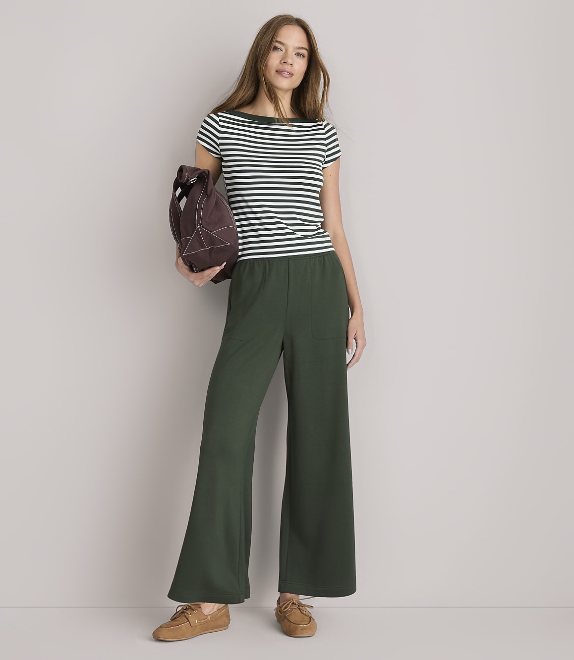A woman in a striped shirt and wide-leg green pants stands against a plain white background, holding a brown bag.