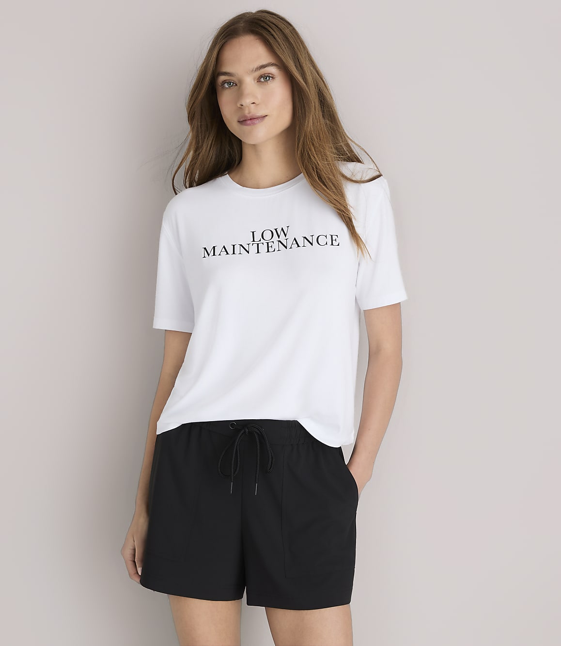 A woman in a white "LOW MAINTENANCE" t-shirt and black baseball cap poses against a plain white background.