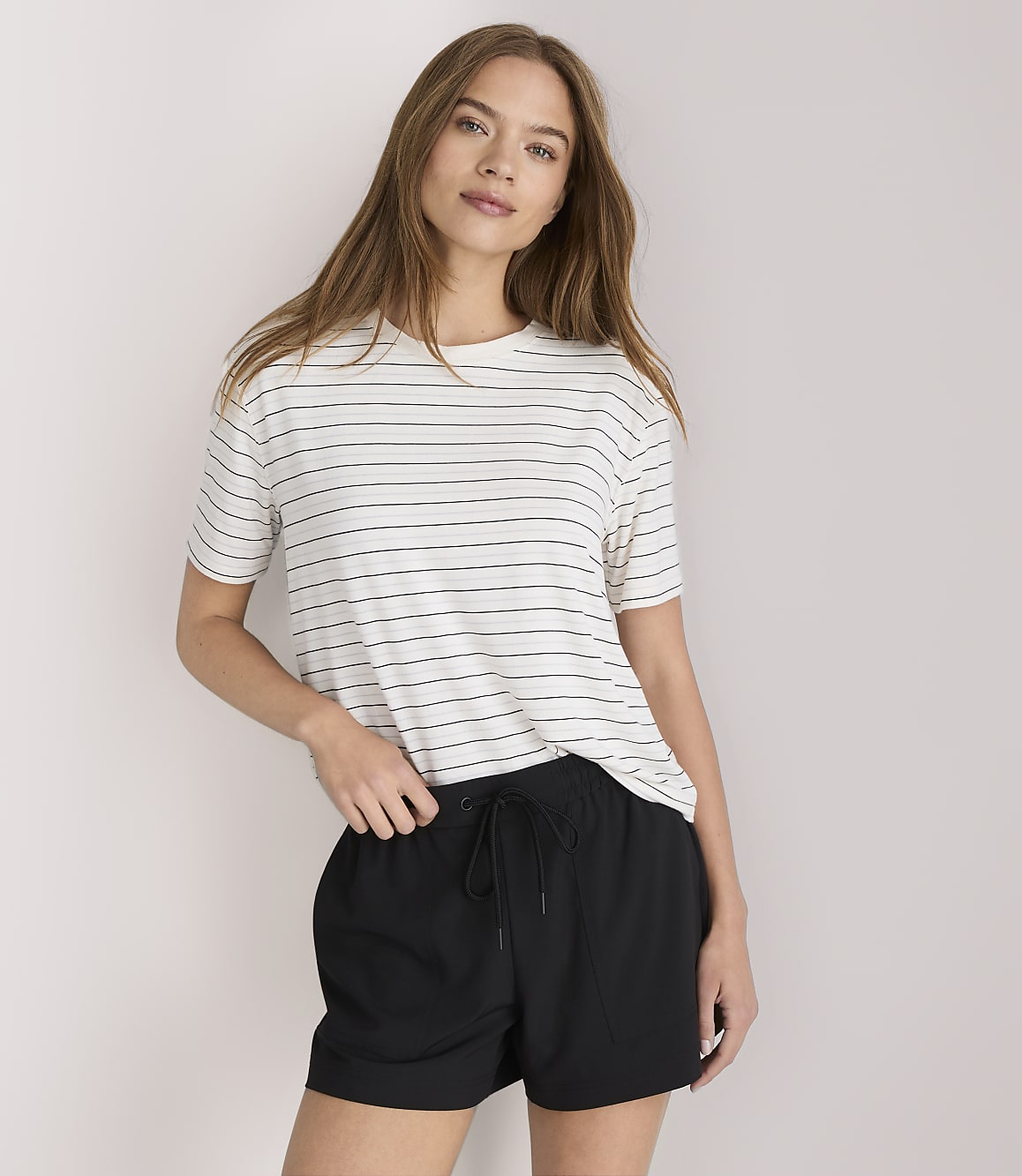A young woman in a striped t-shirt and black shorts stands against a plain white background.