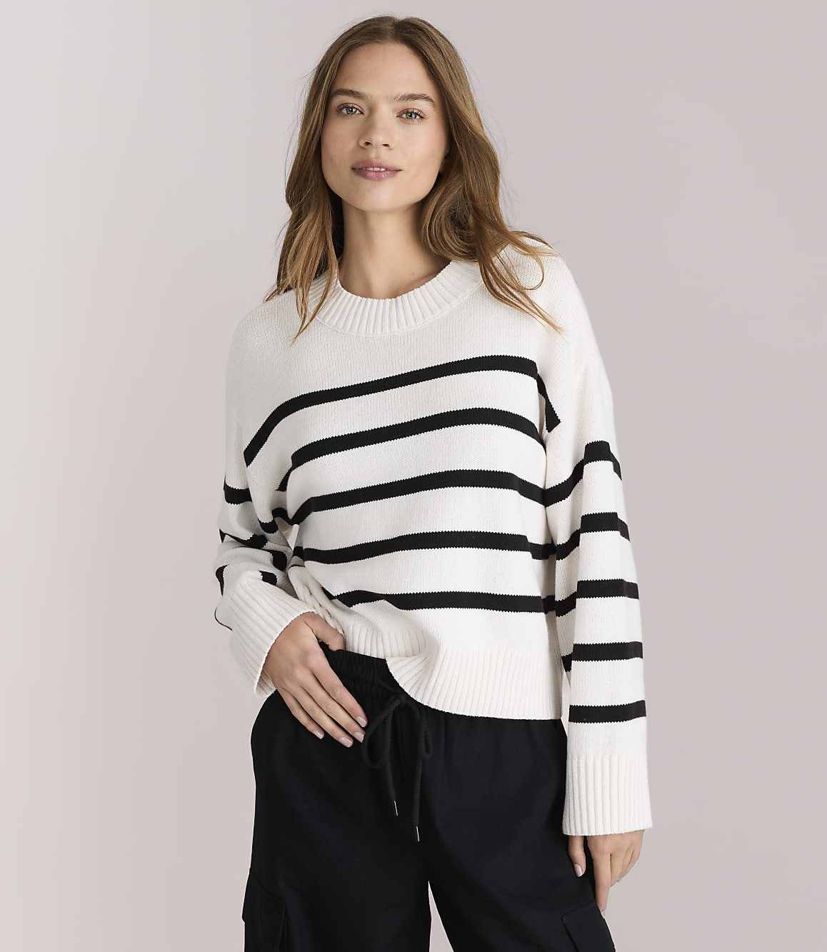 A young woman in a striped sweater and black pants sits cross-legged against a plain white background.