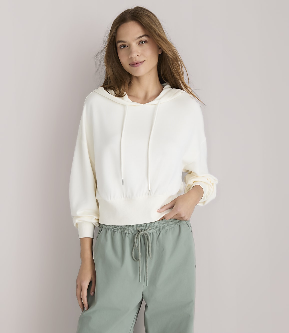 A young woman in a cream hoodie and green drawstring pants stands against a plain white background.