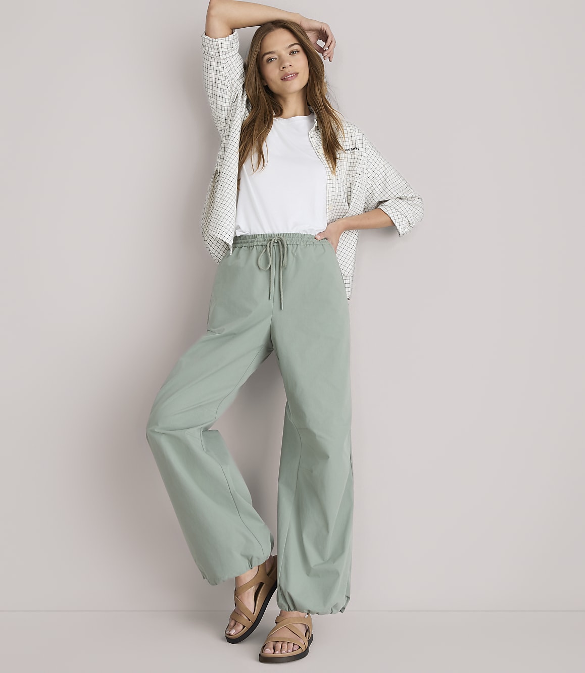 A woman in a white t-shirt, patterned shirt, and loose green pants stands against a plain white background.