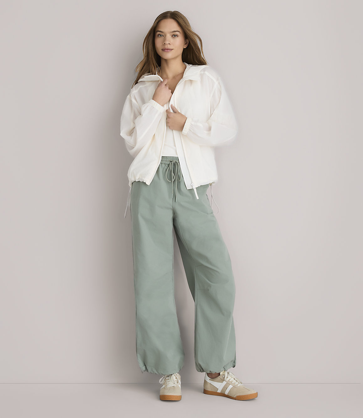 A woman in a white jacket and green pants stands against a plain white background.