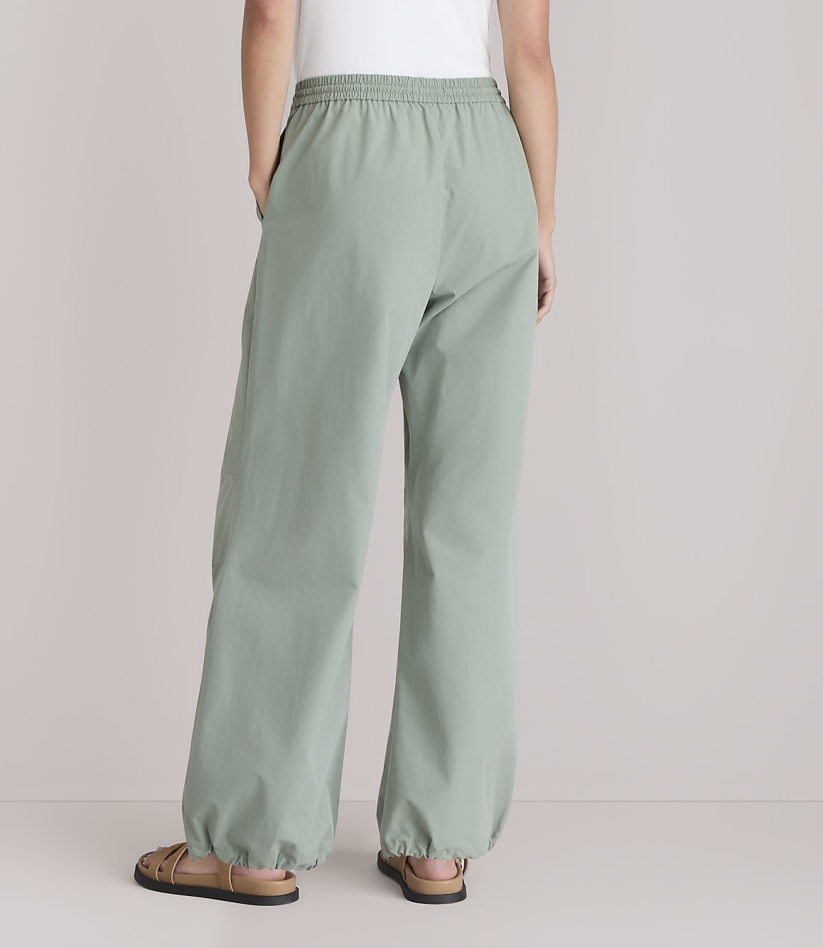 A person wearing loose-fitting, light green pants with elastic cuffs stands with their back to the camera against a plain white background.
