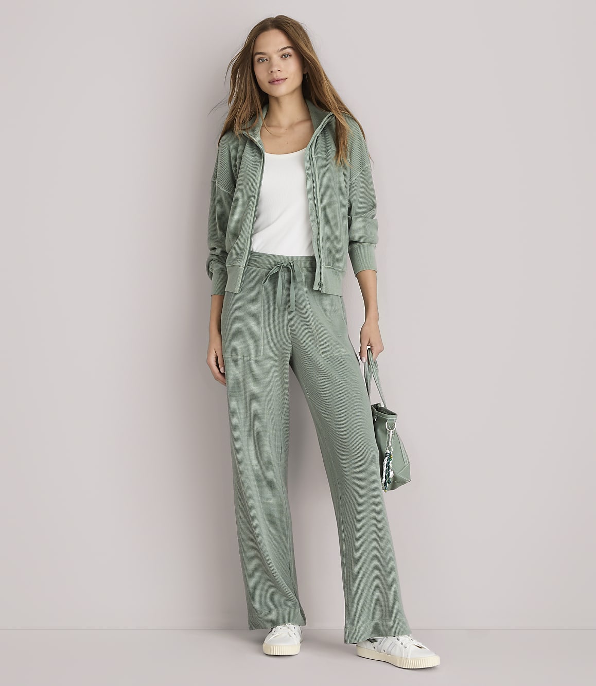 A woman in a sage green loungewear set stands against a plain white background, holding a matching bag.