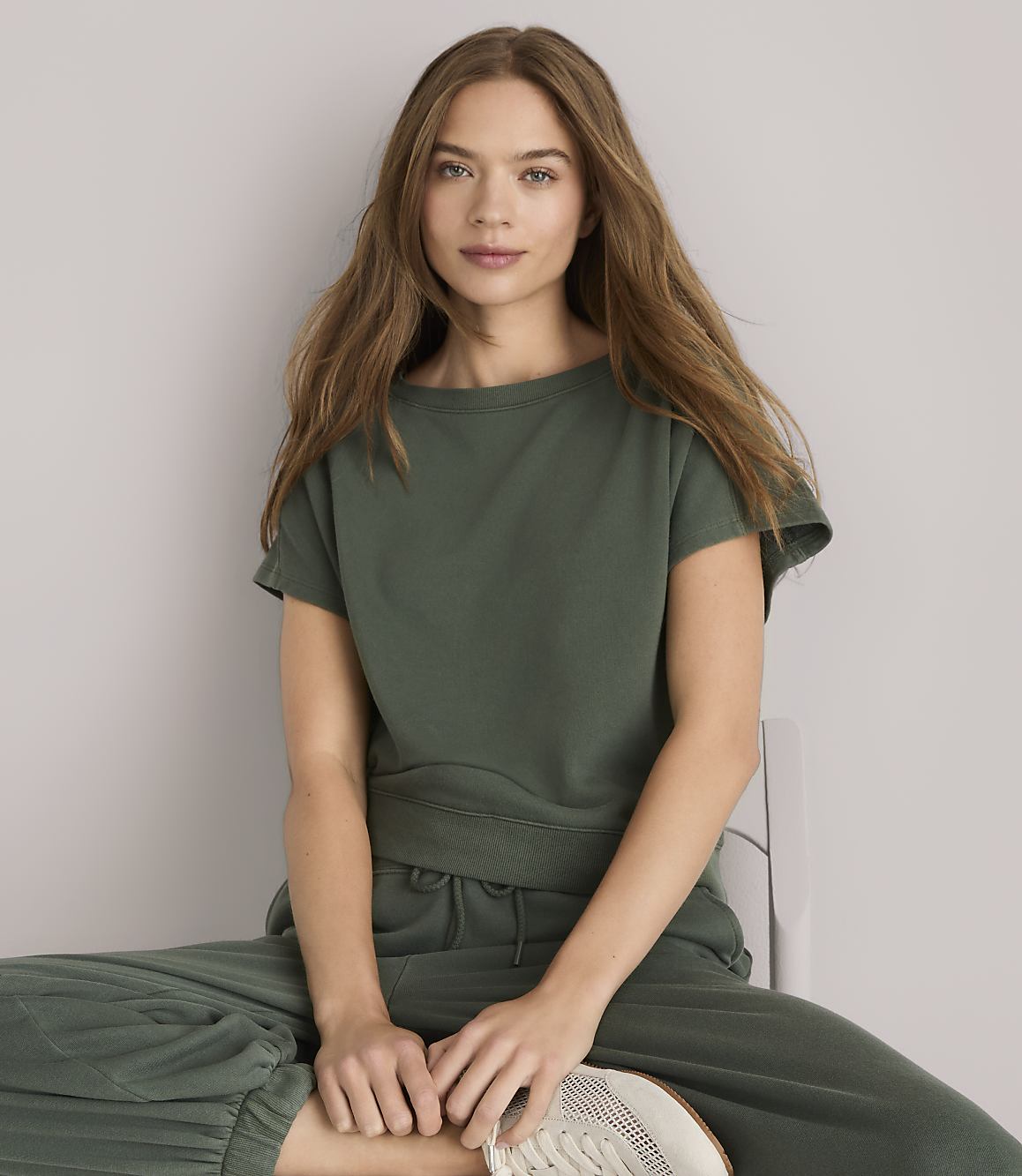 A young woman with long, wavy brown hair, wearing a green t-shirt and pants, sits on a white chair against a plain white background.
