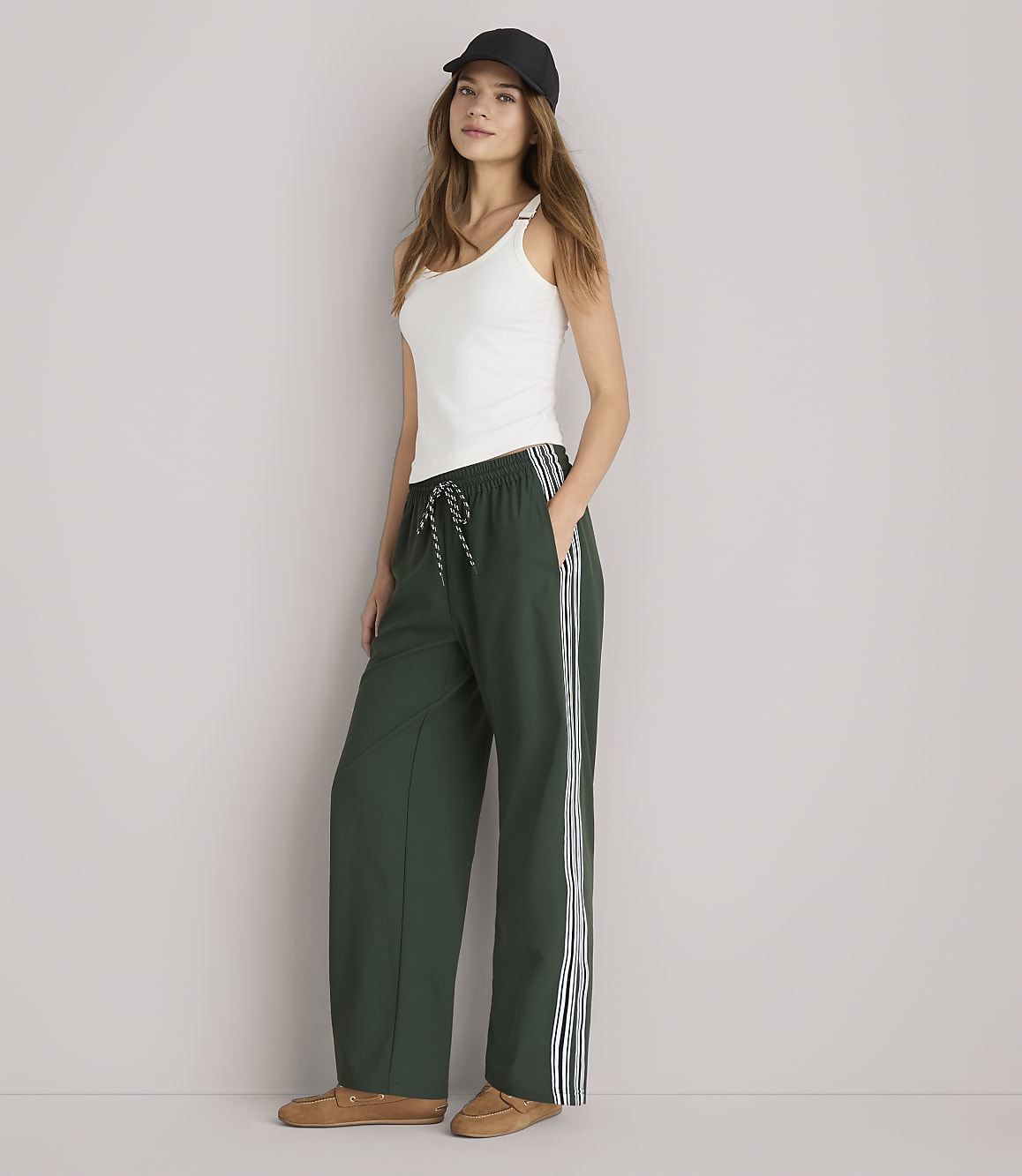 A woman in a white tank top and green track pants stands against a plain white background.
