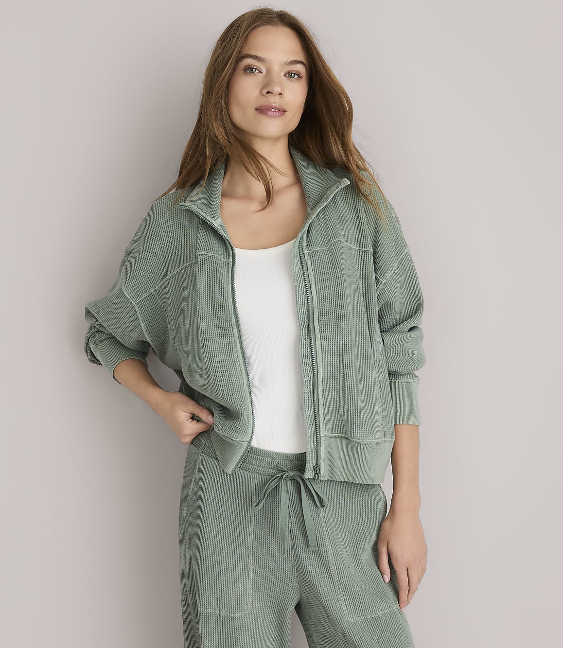 A woman in a green zip-up jacket and matching pants stands against a plain white background.