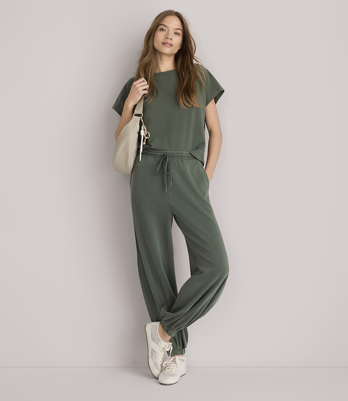 A woman in a green loungewear set and sneakers stands with a cream-colored bag slung over her shoulder against a plain white background.