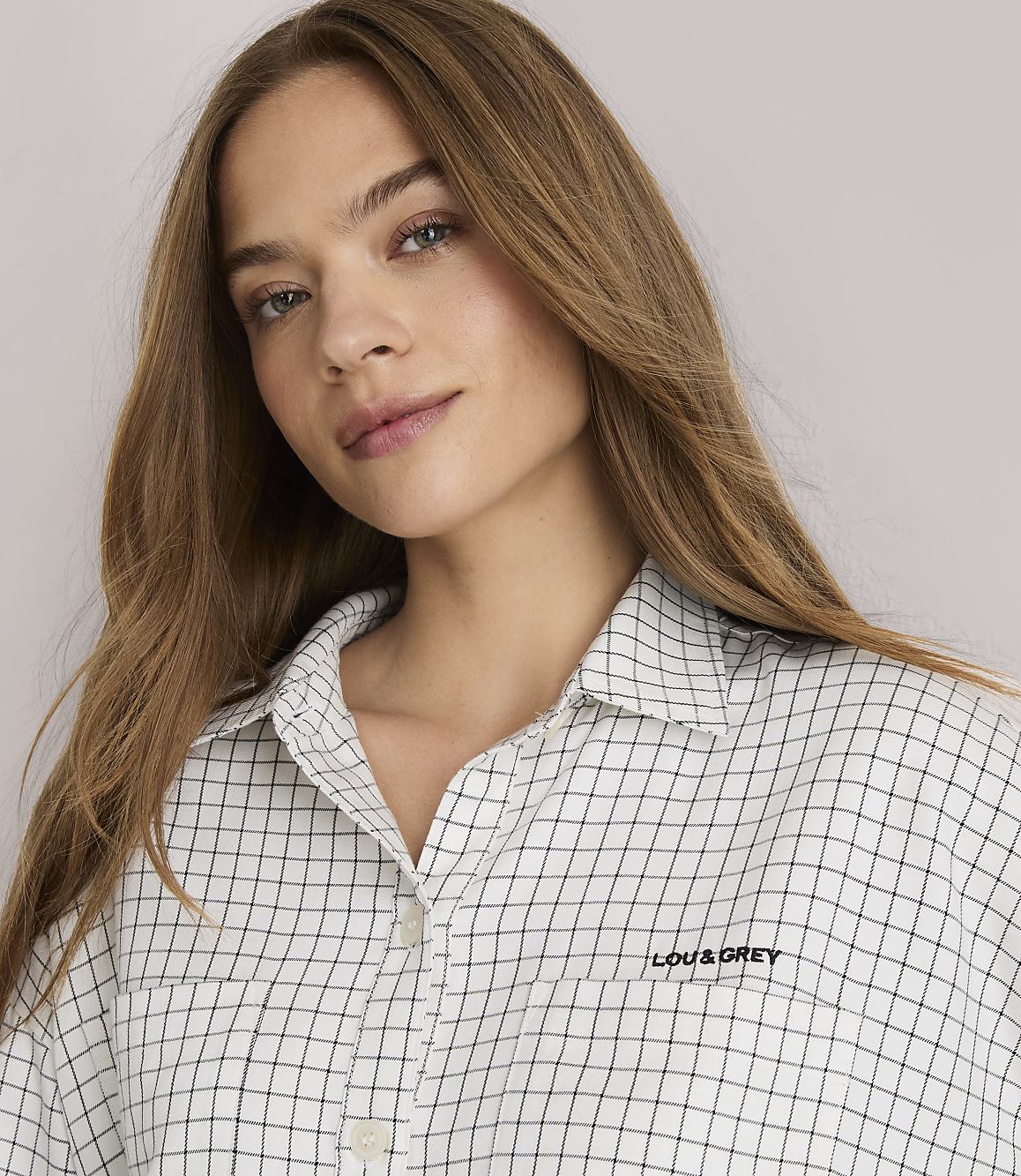 A young woman with long brown hair stands in the foreground wearing a checkered shirt and striped pants, against a plain white background.