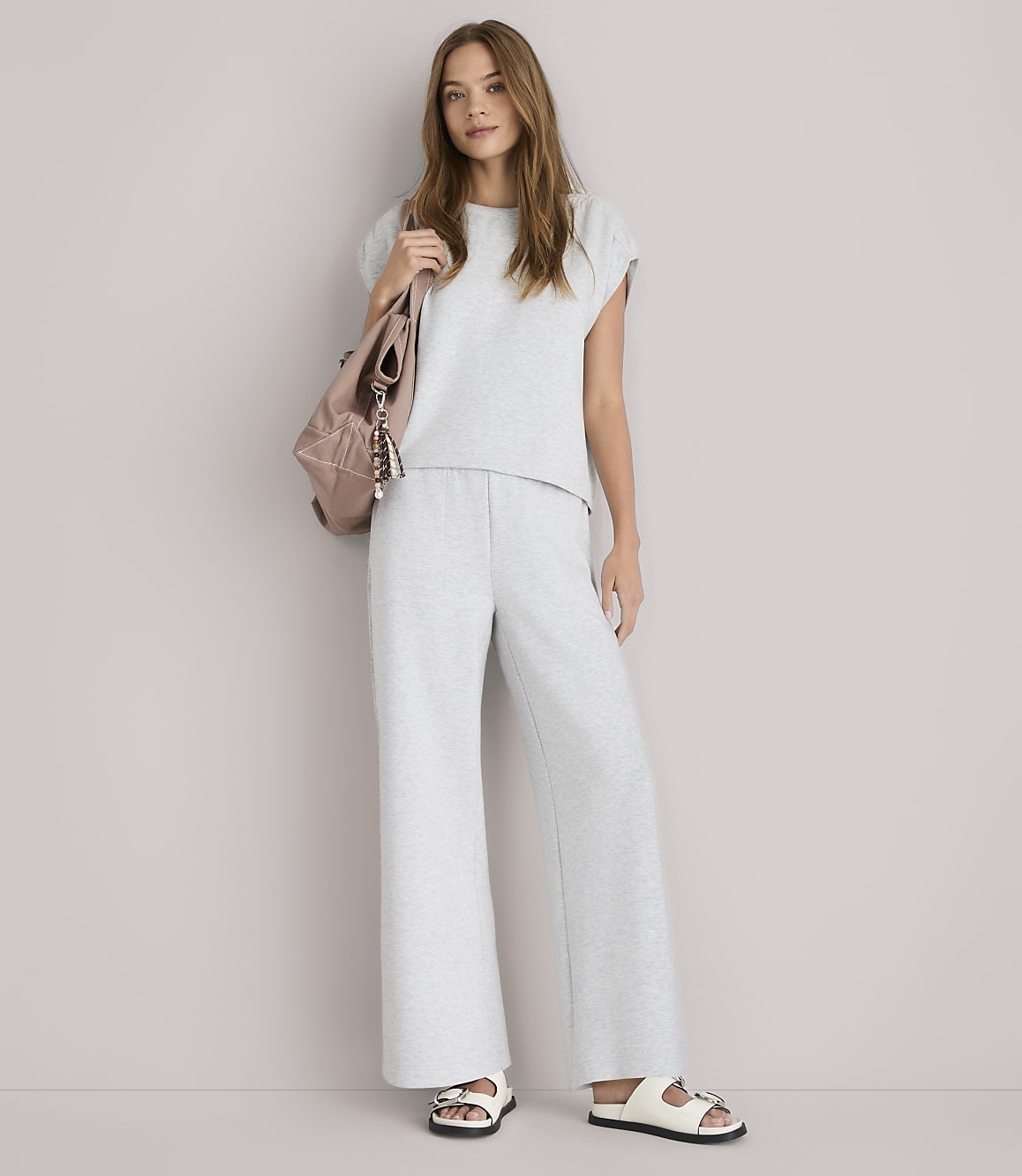 A young woman in a grey loungewear set and white sandals stands against a plain white background, with a pink backpack slung over her shoulder.