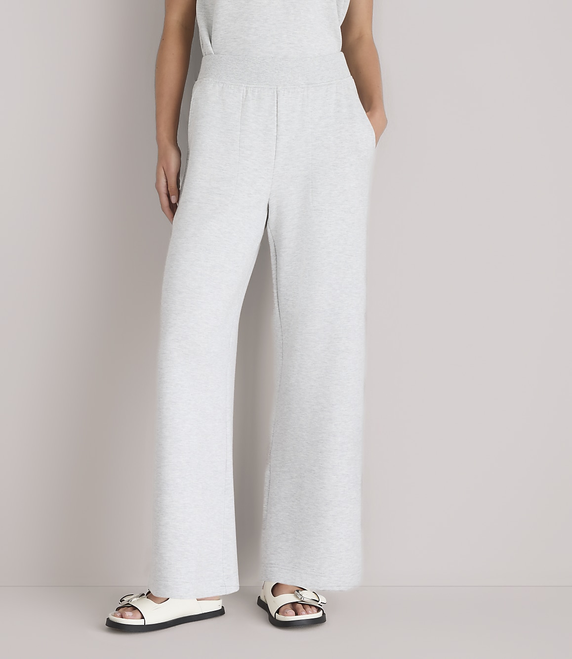 A person wearing light gray wide-leg sweatpants and white sandals stands against a plain white background.