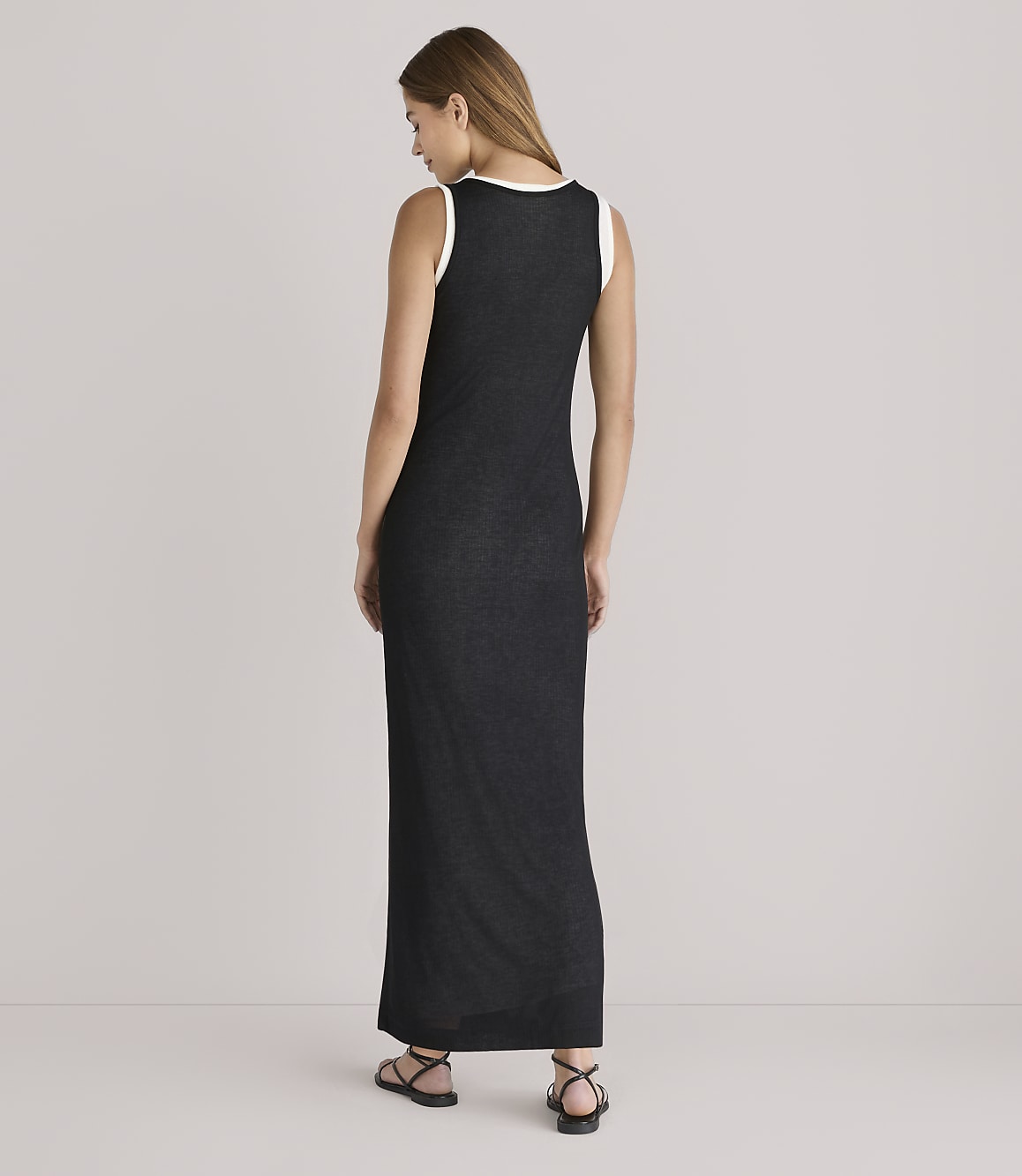 A woman in a black maxi dress with a white trim stands facing away from the camera against a plain, light-colored background.