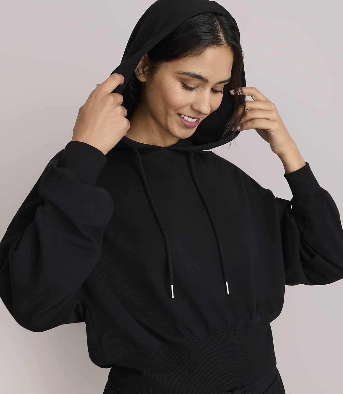 A smiling woman in a black hoodie adjusts the hood against a plain gray background.