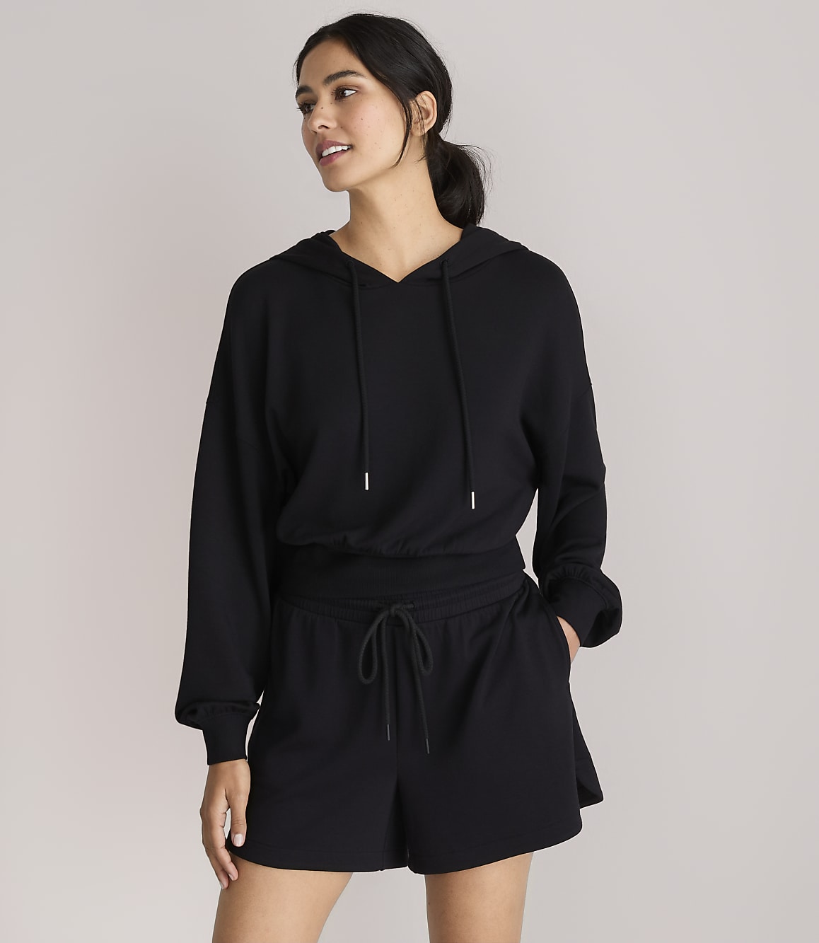 A woman in a black hoodie and shorts stands against a plain white background.