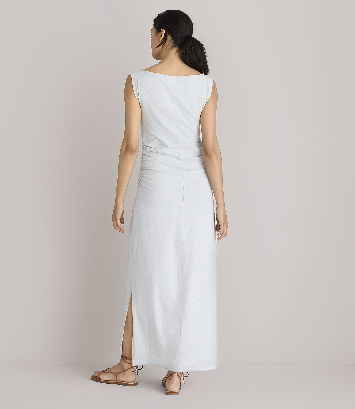 A woman in a light gray maxi dress with ruched detailing and a side slit stands with her back to the camera against a plain white background.