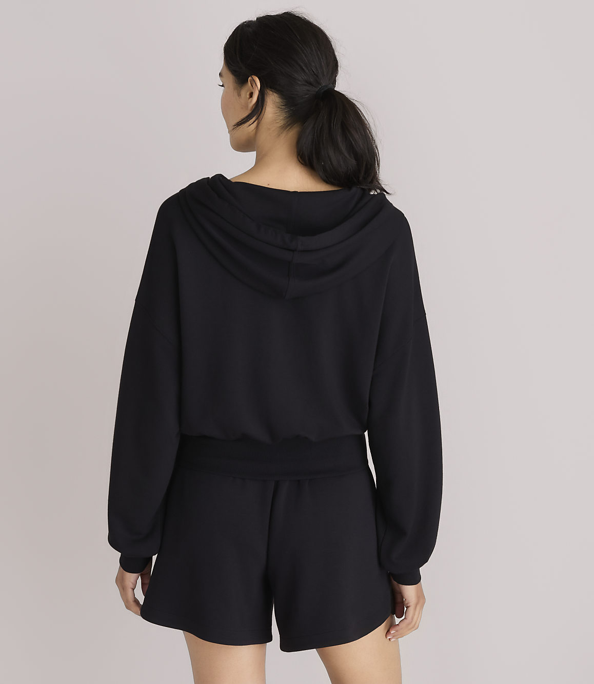 A woman in a black hoodie and shorts stands against a plain white background.